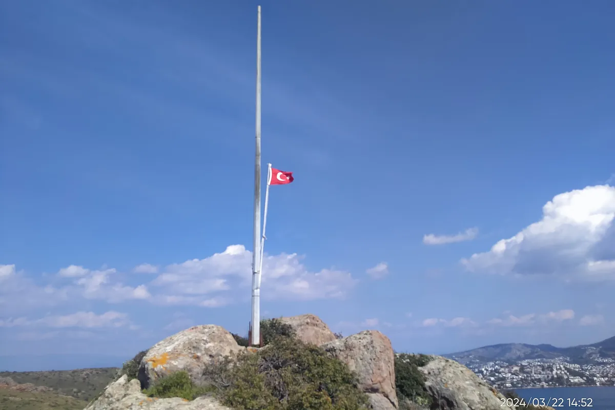 Kayalık bir tepe zirvesinde Türk bayrağı ve arkada geniş bir Ege manzarası